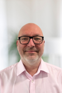Headshot of a smiling bald man with a short grey beard and black-framed rectangular glasses, wearing a light pink button-up shirt. The background is softly blurred in neutral tones with a hint of greenery.