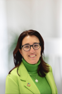 Headshot of a smiling woman with shoulder-length auburn hair and black-framed glasses, wearing a bright green turtleneck and matching lime green blazer. She wears a tree-of-life pendant necklace and an SDG wheel pin on her lapel. The background is softly blurred in neutral tones.