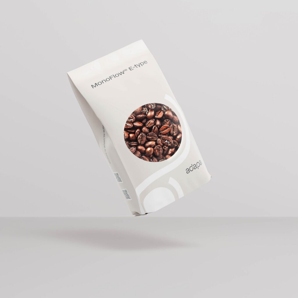 web-optimised-adapa_MonoFlow(re) E-type_coffee A light gray flexible stand-up pouch floating diagonally against a light gray background. The front features the text "MonoFlow^re E-type" near the top and a circular window displaying whole roasted coffee beans inside, with two QR codes on the side gusset and the dark "adapa" brand logo on the lower right.